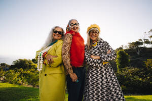 Reife, exzentrische Frauen, die in die Kamera lächeln, während sie zusammen in einem Park stehen. Eine Gruppe selbstbewusster älterer Frauen in farbenfroher Freizeitkleidung. Drei stilvolle ältere Frauen, die ihre goldenen Jahre genießen. - JLPSF09165