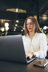 Young businesswoman working on a laptop in a modern workspace. Businesswoman typing on a laptop while listening to music with earphones. Female entrepreneur sitting at a table alone in an office. - JLPSF09221