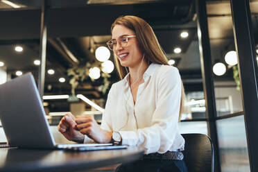 Glückliche junge Geschäftsfrau, die einen Videoanruf in einem modernen Arbeitsraum entgegennimmt. Junge Geschäftsfrau, die während eines virtuellen Meetings an ihrem Laptop lächelt. Fröhliche Unternehmerin, die an einem Tisch in einem Büro sitzt. - JLPSF09224