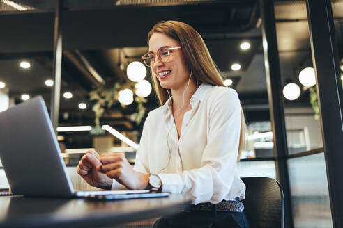 Happy young businesswoman taking a video call in a modern workspace. Young businesswoman smiling during a virtual meeting on her laptop. Cheerful female entrepreneur sitting at a table in an office. - JLPSF09224