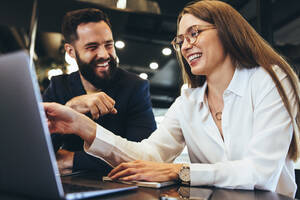 Erfolgreiche Geschäftsleute, die lächelnd mit einem Laptop in einem Büro arbeiten. Glückliche junge Unternehmer, die in einem modernen Arbeitsbereich zusammenarbeiten. Zwei fröhliche Geschäftsleute, die zusammen an einem Tisch sitzen. - JLPSF09232