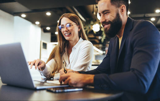 Cheerful businesspeople using a laptop in an office. Happy young entrepreneurs smiling while working together in a modern workspace. Two young businesspeople sitting together at a table. - JLPSF09238