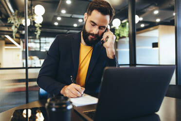 Businessman writing notes during a phone call. Young businessman communicating with his clients while sitting in an office. Focused entrepreneur sitting alone in a modern workspace. - JLPSF09241