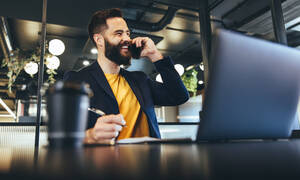 Glücklicher Geschäftsmann, der während eines Telefonats lächelt. Junger Geschäftsmann, der Notizen schreibt, während er mit seinen Kunden in einem Büro kommuniziert. Fröhlicher Unternehmer, der allein in einem modernen Arbeitsbereich sitzt. - JLPSF09242