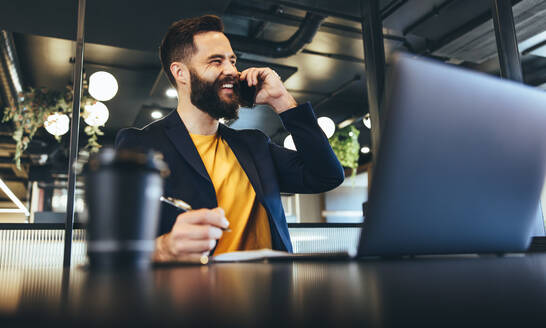 Happy businessman smiling during a phone call. Young businessman writing notes while communicating with his clients in an office. Cheerful entrepreneur sitting alone in a modern workspace. - JLPSF09242