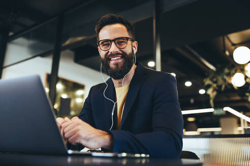 Erfolgreicher Unternehmer, der in einem Büro in die Kamera lächelt. Glücklicher junger Geschäftsmann, der Musik hört, während er an einem Laptop arbeitet. Fröhlicher Geschäftsmann, der allein in einem modernen Arbeitsraum sitzt. - JLPSF09246