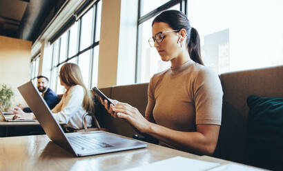 Businesswoman reading a text message in a co-working space. Female entrepreneur using a smartphone while wearing wireless earphones. Young businesswoman sitting in a modern workspace. - JLPSF09248