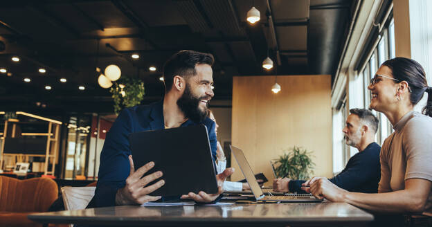 Mitarbeiter lächeln sich an, während sie an einem Tisch in einem Co-Working Space sitzen. Zwei glückliche Geschäftsleute teilen sich einen Arbeitsplatz in einem Großraumbüro. Fröhliche Jungunternehmer, die drahtlose Technologie nutzen. - JLPSF09252