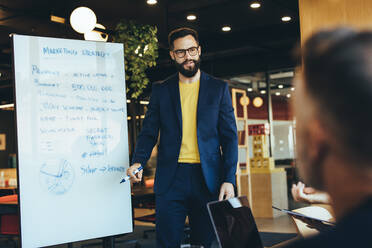 Business manager giving a presentation during a meeting with his team. Confident young businessman discussing his marketing strategy with his colleagues in a modern workspace. - JLPSF09265