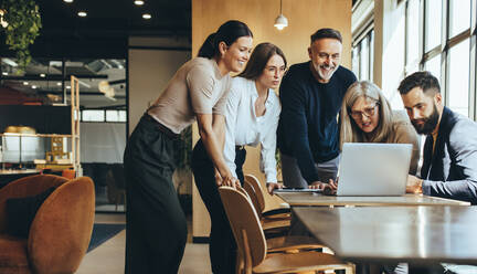 Eine Gruppe von Geschäftsleuten arbeitet mit einem Laptop an einem neuen Projekt. Ein Team von glücklichen Geschäftsleuten schaut auf den Laptop-Bildschirm, während sie in einem modernen Arbeitsraum zusammenarbeiten. - JLPSF09275