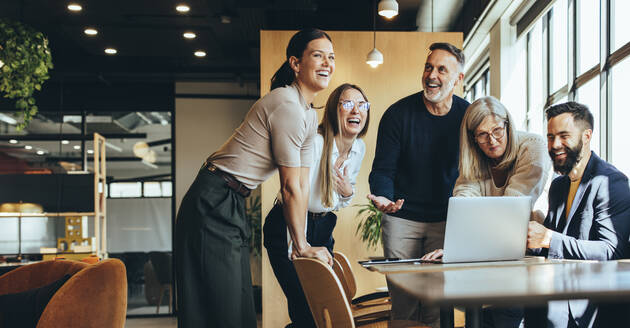 Fröhliche Geschäftsleute lachen, während sie in einem Büro an einem neuen Projekt arbeiten. Eine Gruppe verschiedener Geschäftsleute benutzt einen Laptop, während sie in einem modernen Arbeitsraum zusammenarbeiten. - JLPSF09277