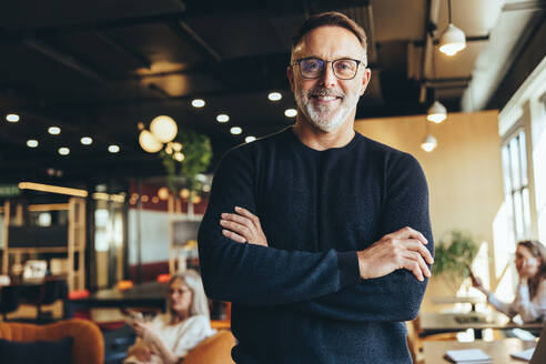 Mature businessman standing in a co-working space. Happy mature businessman smiling at the camera while standing with his arms crossed. Experienced entrepreneur working in a modern workspace. - JLPSF09281