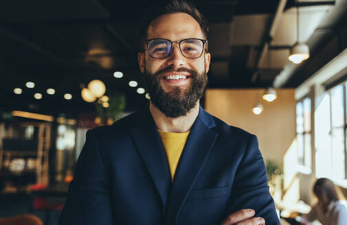 Fröhlicher Geschäftsmann, der in einem Co-Working Space in die Kamera lächelt. Glücklicher junger Geschäftsmann, der mit verschränkten Armen alleine steht. Erfolgreicher junger Unternehmer, der in einem modernen Arbeitsraum arbeitet. - JLPSF09289