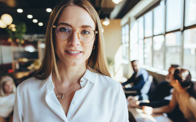 Fröhliche Geschäftsfrau, die in einem Co-Working Space in die Kamera lächelt. Nahaufnahme einer glücklichen jungen Unternehmerin, die an einem modernen Arbeitsplatz steht, mit ihren Arbeitskollegen im Hintergrund. - JLPSF09292