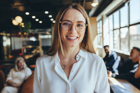Erfolgreiche junge Geschäftsfrau, die in einem Co-Working Space in die Kamera lächelt. Glückliche junge Unternehmerin an einem modernen Arbeitsplatz mit ihren Mitarbeitern im Hintergrund. - JLPSF09293