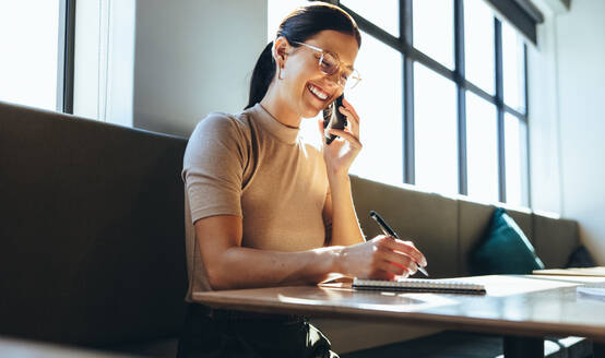 Unternehmerin, die während eines Telefonats Notizen schreibt. Glückliche junge Geschäftsfrau, die mit ihren Geschäftspartnern Pläne schmiedet. Geschäftsfrau, die lächelt, während sie in einem modernen Arbeitsraum arbeitet. - JLPSF09294