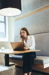 Female business professional speaking on a phone call. Young businesswoman using a laptop while making plans with her business partners. Female entrepreneur working in a modern workspace. - JLPSF09323