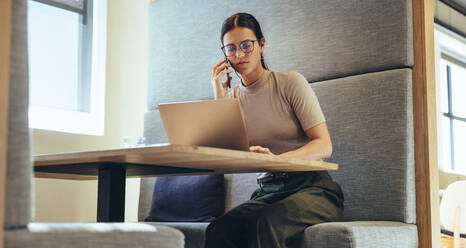 Entrepreneur speaking on the phone in a modern workspace. Young businesswoman using a laptop during a phone call with her business partners. Female business professional making business plans. - JLPSF09329