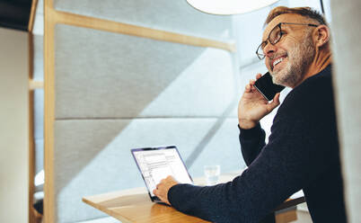 Experienced entrepreneur smiling during a phone call. Happy mature businessman communicating with his partners over the phone. Mature business professional working in a modern workspace. - JLPSF09332