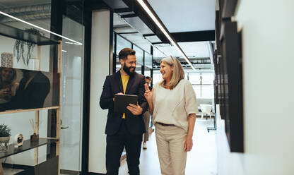 Unterschiedliche Geschäftsleute, die während einer Diskussion in einem Büro lachen. Zwei glückliche Geschäftskollegen, die ein digitales Tablet benutzen, während sie zusammen in einem modernen Arbeitsraum spazieren gehen. - JLPSF09340