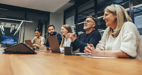 Fröhliche Geschäftsleute, die während einer Besprechung lachen. Eine Gruppe glücklicher Geschäftsleute, die die Zusammenarbeit an einem modernen Arbeitsplatz genießen. Ein Team verschiedener Kollegen bei einer Besprechung in einem Sitzungssaal. - JLPSF09354