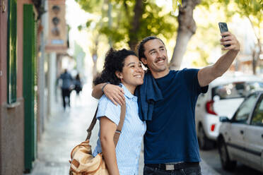 Lächelnder Mann mit Arm um Freundin spricht Selfie auf Handy - JOSEF14469