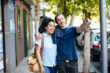 Smiling man with arm around of girlfriend pointing and walking on footpath - JOSEF14472