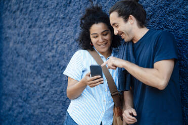 Smiling young man pointing at girlfriend using smart phone in front of wall - JOSEF14479