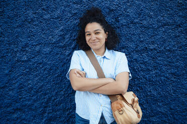 Smiling woman wearing shoulder bag with arms crossed in front of blue wall - JOSEF14485