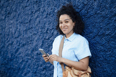 Lächelnde Frau mit Umhängetasche, die ihr Smartphone vor einer blau strukturierten Wand hält - JOSEF14486