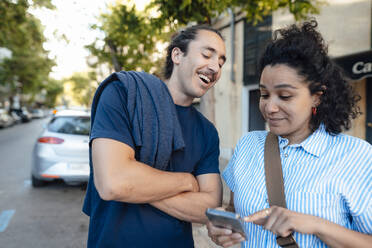 Happy man looking at girlfriend using mobile phone on street - JOSEF14488