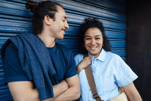 Lächelnde Frau neben ihrem Freund mit verschränkten Armen vor einer blauen Wand - JOSEF14498