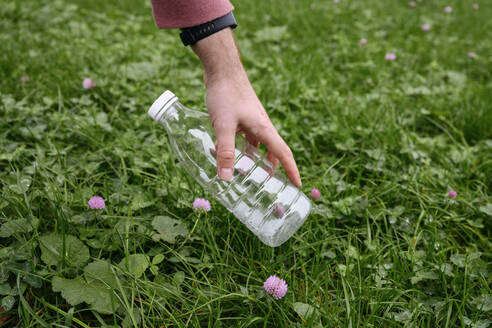Hand eines Mannes, der eine Plastikflasche aus dem Gras sammelt - EYAF02196