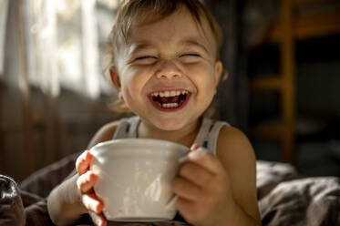 Cute baby with cup laughing in bed at home - ANAF00182