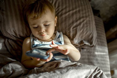 Niedliches Kleinkind mit Smartphone im Bett liegend im Schlafzimmer - ANAF00185