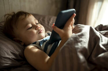 Cute baby boy using smart phone in duvet in bed at home - ANAF00187