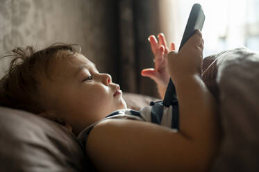 Kleiner Junge mit Mobiltelefon im Bett liegend - ANAF00188