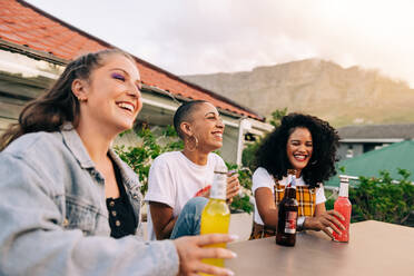 Unbekümmerte Freundinnen genießen ein paar kalte Biere auf einem Dach. Drei glückliche Freundinnen lachen fröhlich, während sie zusammen abhängen. Eine Gruppe von Freunden, die eine gute Zeit am Wochenende haben. - JLPSF09365