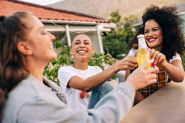 Drei Freundinnen stoßen auf einer Dachterrasse auf ihre Freundschaft an. Drei Freundinnen lächeln fröhlich und heben ihre Bierflaschen. Glückliche Freundinnen, die am Wochenende zusammen abhängen. - JLPSF09366