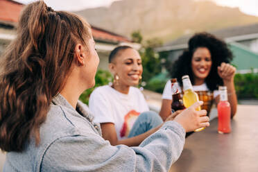 Fröhliche junge Frau stößt mit ihren Freunden auf einem Dach auf die Freundschaft an. Eine Gruppe von drei fröhlichen Freundinnen genießt ein paar kalte Biere, während sie am Wochenende zusammen abhängen. - JLPSF09367