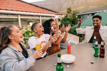 Friends cheering happily while having some cold beers and popcorn. Group of carefree young people hanging out together on a rooftop. Cheerful friends having a good time together on the weekend. - JLPSF09369