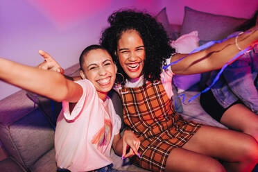 Zwei glückliche junge Frauen machen ein Selfie auf einer Hausparty. Zwei glückliche junge Frauen machen ein gemeinsames Foto, während sie auf einer Couch im Neonlicht sitzen. Beste Freundinnen, die am Wochenende eine gute Zeit miteinander verbringen. - JLPSF09378