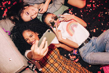 Niedliche Selfies nur. Hoher Blickwinkel von drei glücklichen Freunden, die ein Selfie machen, während sie auf dem Boden bei einer Hausparty liegen. Gruppe von fröhlichen weiblichen Freunden, die am Wochenende zusammen Spaß haben. - JLPSF09408