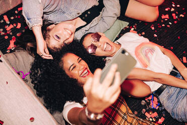 Taking crazy overhead selfies. Top view of three happy friends taking a selfie while lying on the floor at a house party. Group of cheerful female friends having fun together on the weekend. - JLPSF09409