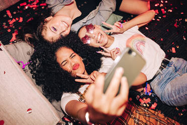 Aufnahme von verrückten Partymomenten. Draufsicht auf drei glückliche Freunde, die ein Selfie machen, während sie auf dem Boden einer Hausparty liegen. Gruppe von fröhlichen Freundinnen, die am Wochenende zusammen Spaß haben. - JLPSF09410