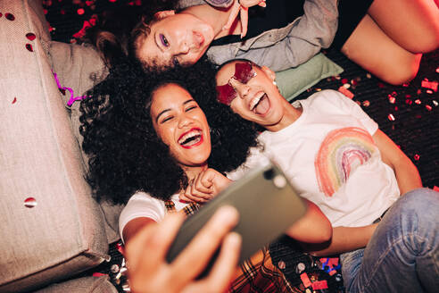 Lebendige Selfies für lebhafte Menschen. Draufsicht auf drei glückliche Freundinnen, die ein Selfie machen, während sie auf dem Boden einer Hausparty liegen. Gruppe von fröhlichen Freundinnen, die am Wochenende zusammen Spaß haben. - JLPSF09411