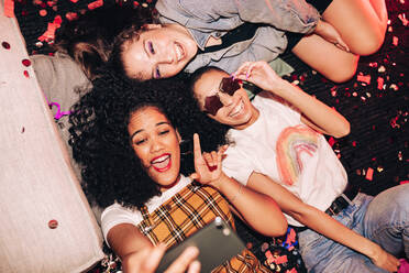 Erinnerungen mit verrückten Selfies machen. Draufsicht auf drei glückliche Freunde, die ein Selfie machen, während sie auf dem Boden einer Hausparty liegen. Gruppe von fröhlichen Freundinnen, die am Wochenende zusammen Spaß haben. - JLPSF09412