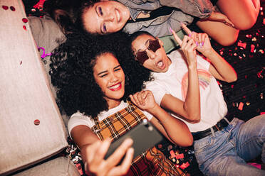 Selfies bei einem Mädelsabend. Drei glückliche Freundinnen machen ein Selfie, während sie auf dem Boden einer Hausparty liegen. Eine Gruppe fröhlicher Freundinnen, die am Wochenende zusammen Spaß haben. - JLPSF09413