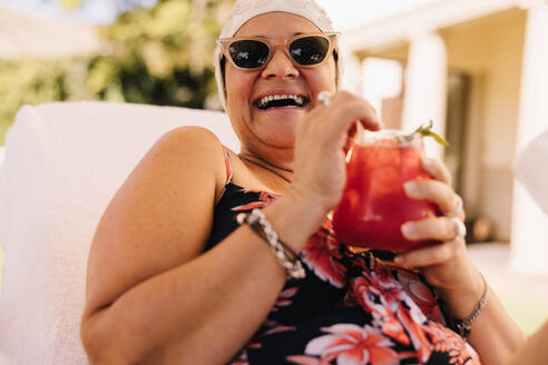 Glückliche ältere Frau, die einen Cocktail im Sommerurlaub genießt. Unbeschwerte ältere Frau, die fröhlich lacht, während sie sich auf einer Liege entspannt. Reife Frau, die sich nach der Pensionierung amüsiert. - JLPSF09433