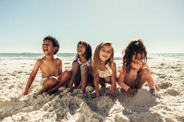 Vier junge Freunde, die fröhlich lachen, während sie am Strand mit Sand spielen. Eine Gruppe liebenswerter kleiner Kinder, die in den Sommerferien eine gute Zeit miteinander verbringen. - JLPSF09453